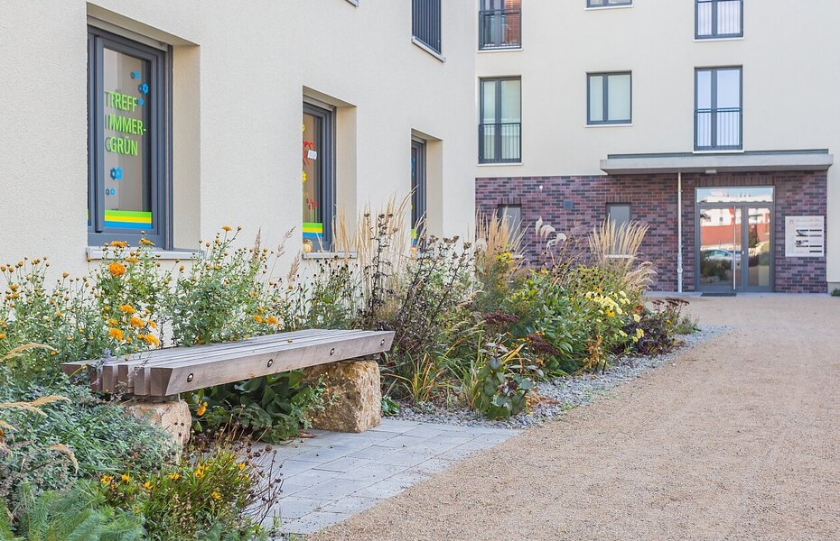 Eingangsbereich der Gartenstadt 2.0 – Quartier Immergrün in Jena mit Sitzbank, Bepflanzung und mehrgeschossigem Wohngebäude